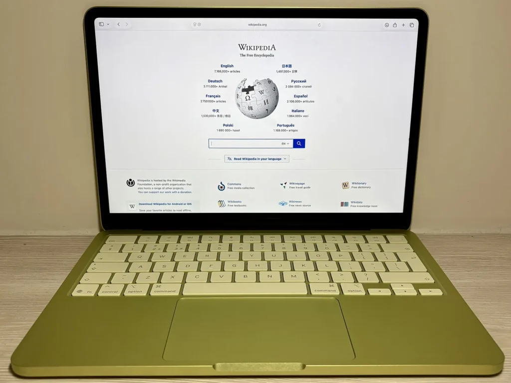 MacBook Neo màu Citrus đặt trên bàn gỗ, màn hình mở trang chủ Wikipedia